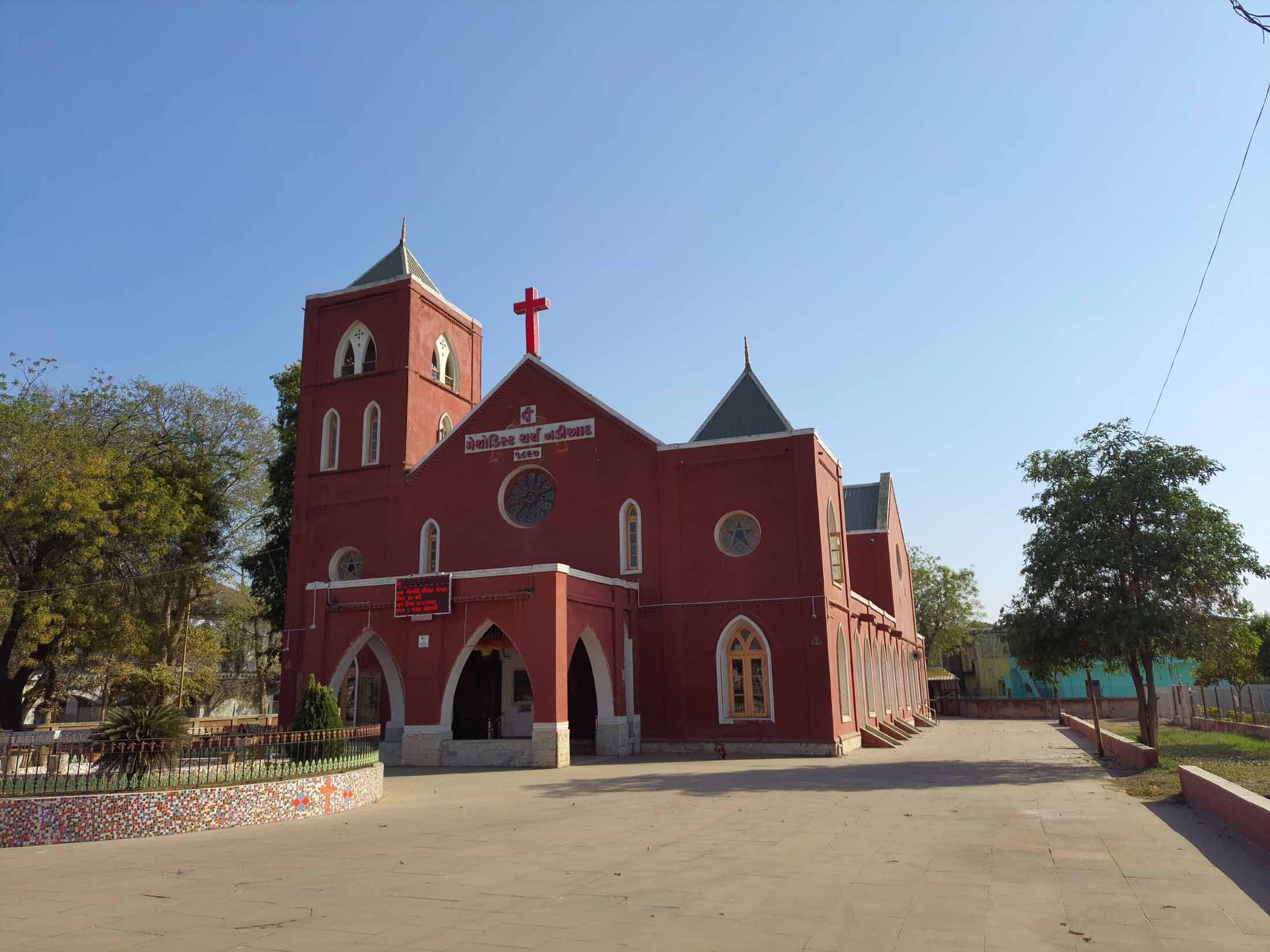 Front elevation of Methodist Church Nadiad, showing the bell tower, red cross and the Gujarati nameplate dated 1897, seen across the paved forecourt.