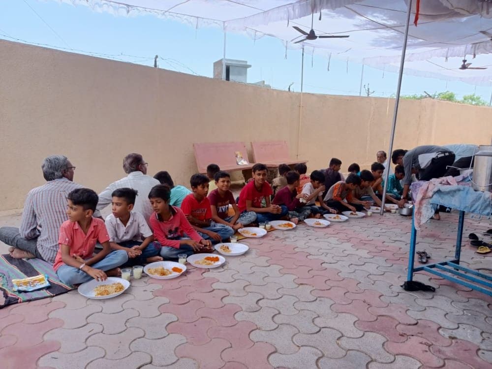 Children and elders seated in a row for a communal meal served under an awning at the church.
