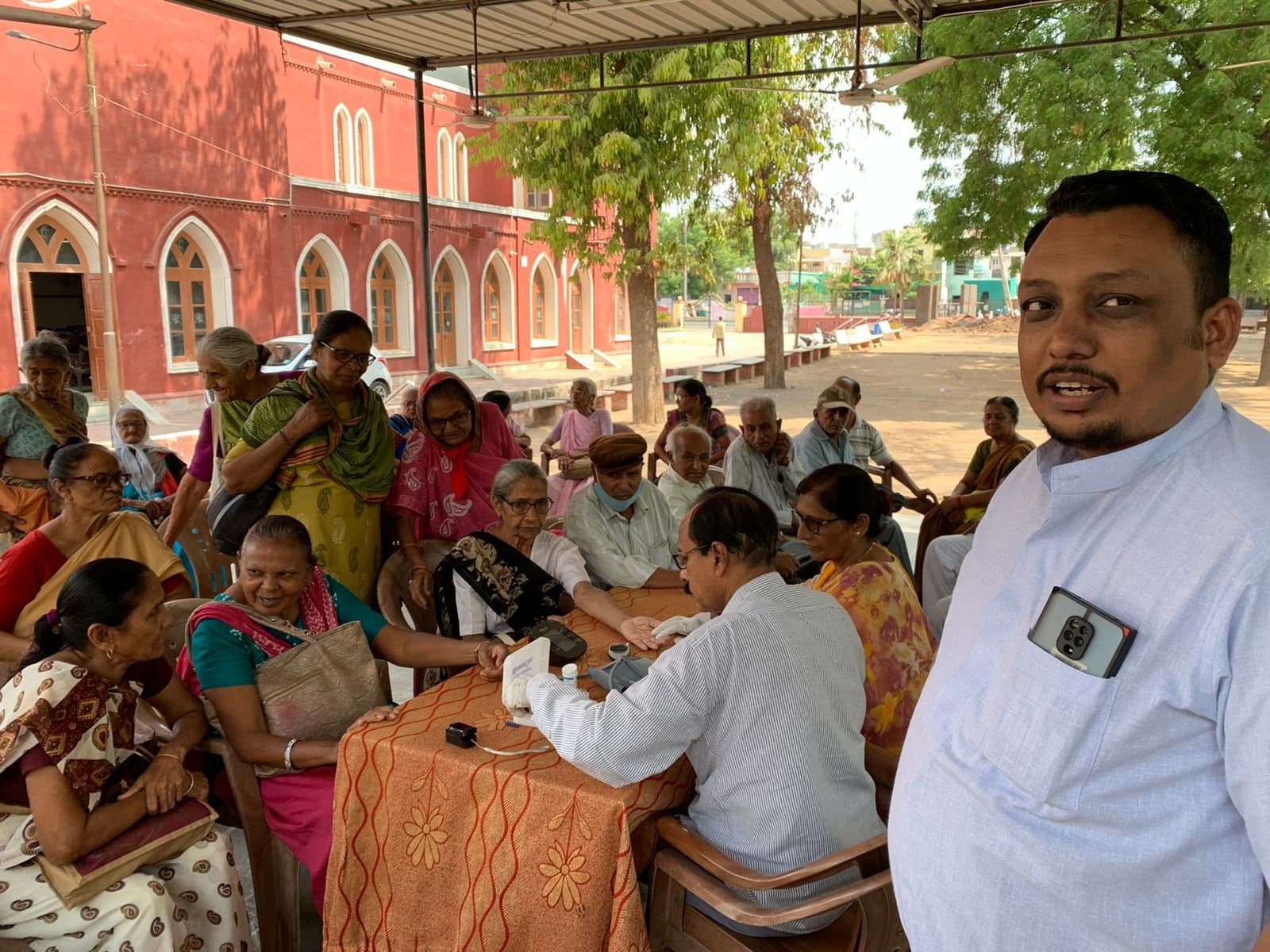 A health check-up camp in the church grounds, with a doctor taking blood pressure readings for older members of the community.